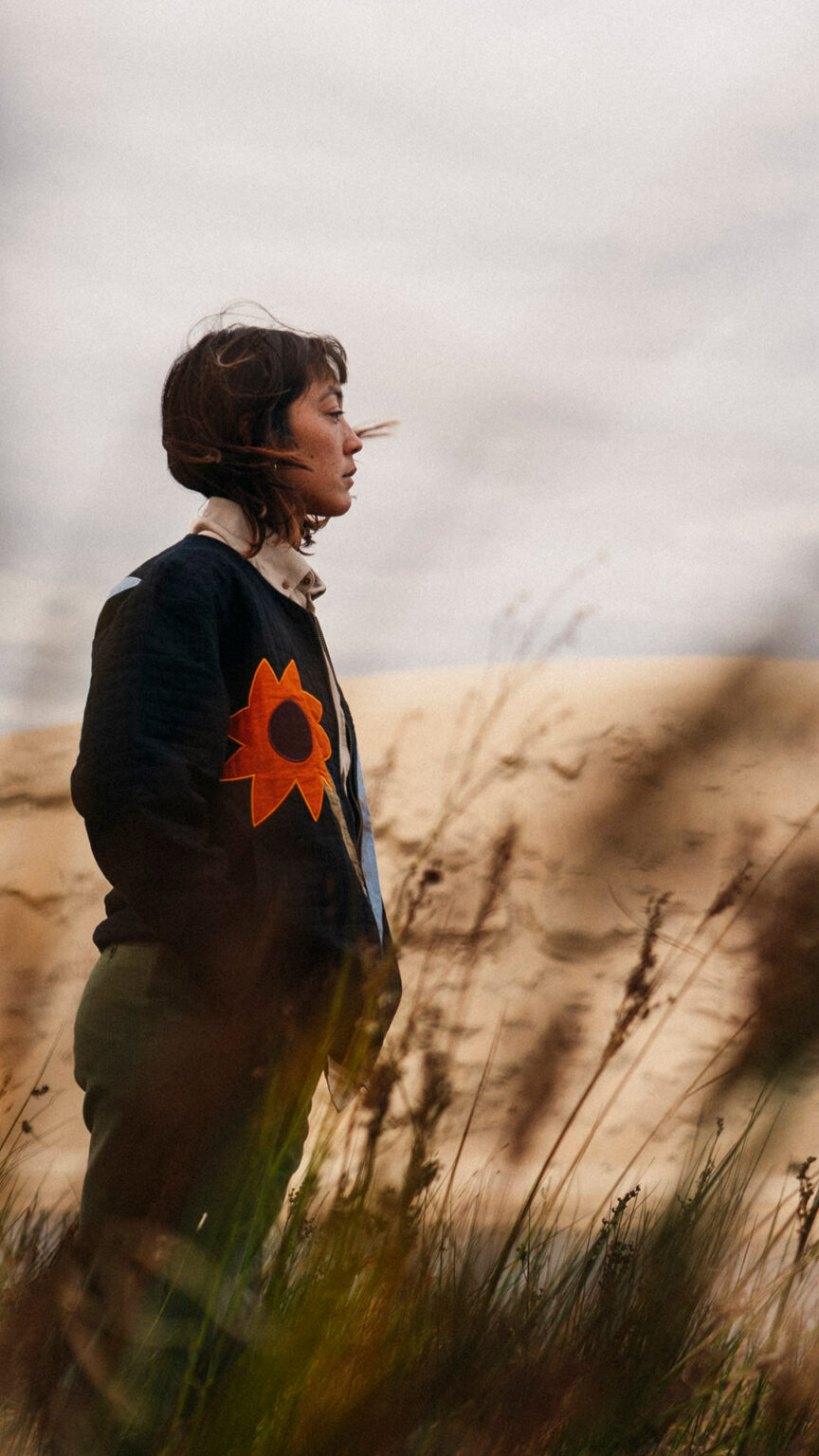 Personne portant le Carbon Black Kigo Zipped Sweatshirt de la marque OLOW dans un paysage naturel de dunes et d’herbes sauvages, vêtement au design graphique et éthique. Person wearing the OLOW Carbon Black Kigo Zipped Sweatshirt in a natural landscape of dunes and wild grass, featuring a graphic, ethical design.